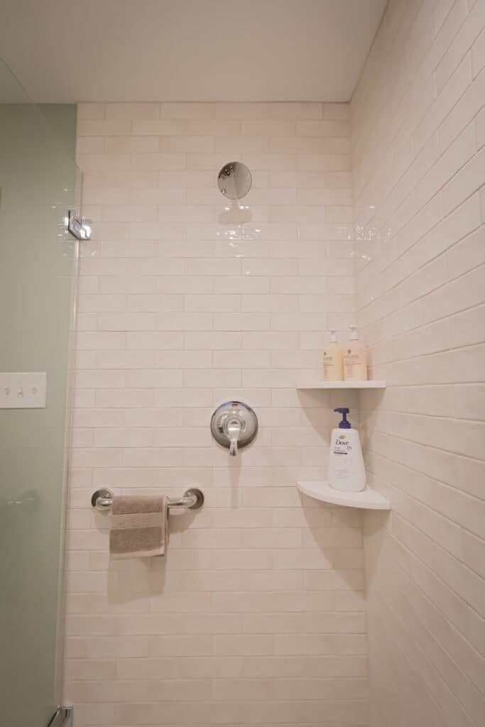 A shower with beige subway tile walls, a metal shower handle, a grab bar holding a folded towel, and two corner shelves with bottles of soap and hair products.