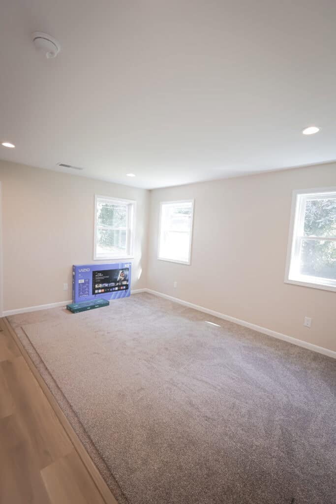 A bright, empty room with beige walls, carpeted flooring, two windows, and a flat-screen TV in a box placed near one wall. Natural light is streaming in through the windows.