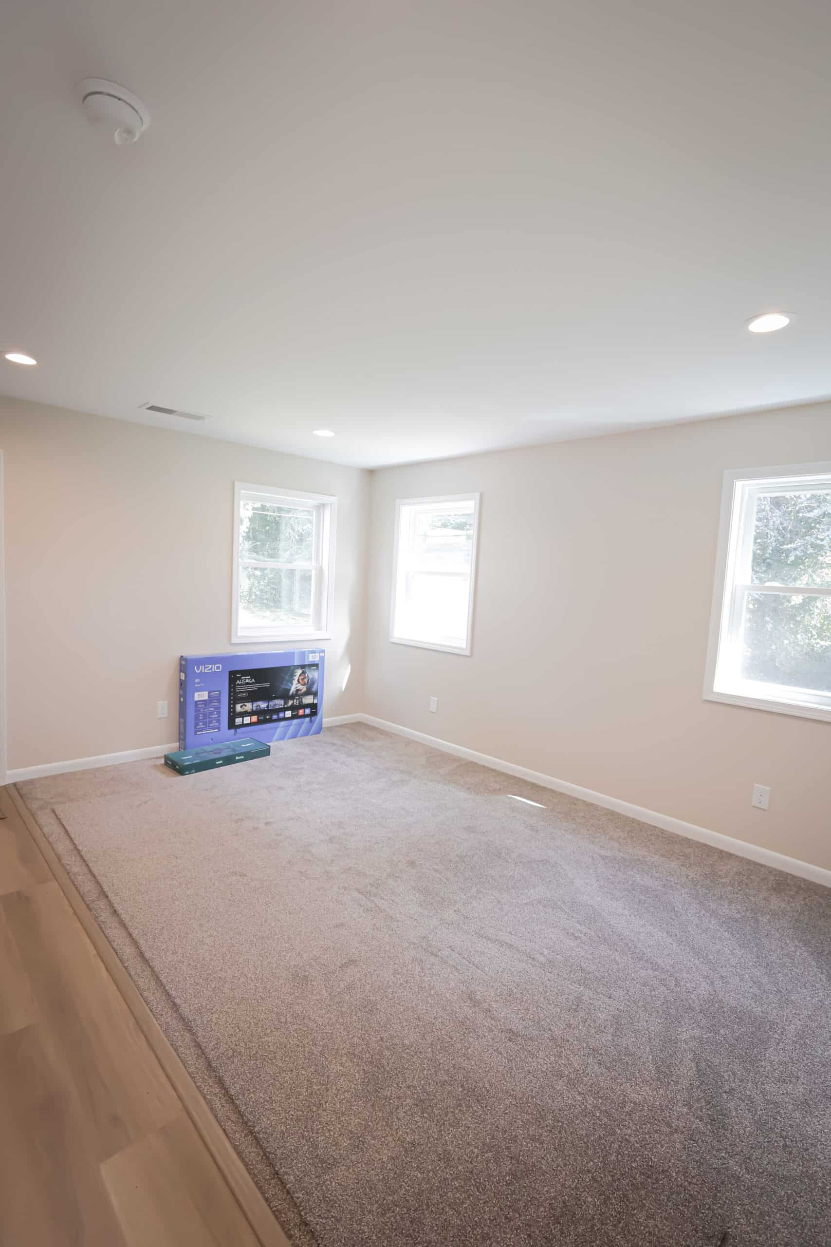 A bright, empty room with beige walls, carpeted flooring, two windows, and a flat-screen TV in a box placed near one wall. Natural light is streaming in through the windows.