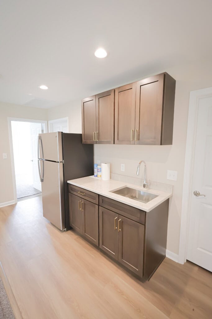 Modern kitchenette with brown cabinets, stainless steel refrigerator, single bowl sink, and a roll of paper towels on the counter. The room has light wood flooring and neutral-colored walls.