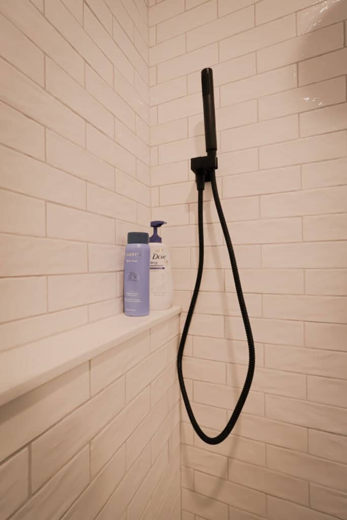A shower with white subway tile walls, a black handheld showerhead with hose, and two bottles—one purple and one white—on a built-in shelf.