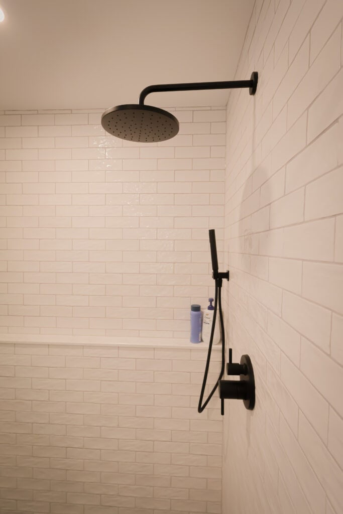 A modern shower with white subway tile walls, a matte black rainfall showerhead, handheld shower, and a built-in shelf holding a blue and white bottle.