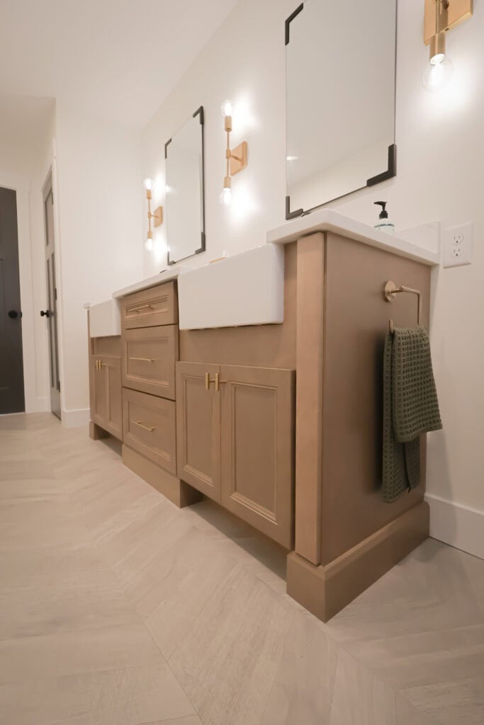 Modern bathroom with double sinks, light wood vanity, gold hardware, two square mirrors, wall-mounted light fixtures, and a green hand towel hanging on the right. The floor features light herringbone tiles.