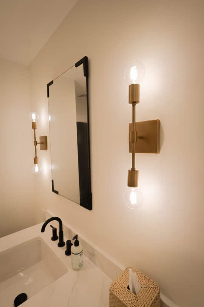 Modern bathroom with a rectangular mirror, two brass wall sconces with exposed bulbs on either side, a white sink with black fixtures, a soap dispenser, and a woven tissue box on a marble countertop.