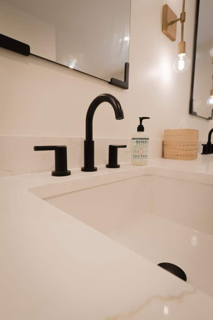 A modern bathroom sink with a matte black faucet, a bottle of Mrs. Meyers hand soap, a woven basket, a large mirror, and a wall-mounted light fixture with an exposed bulb.