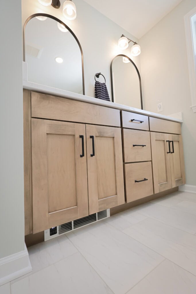 Modern bathroom vanity with light wood cabinets, black handles, double sinks, quartz countertop, two arched mirrors, wall-mounted lights, and white tiled floor. A dark hand towel is hanging between the mirrors.