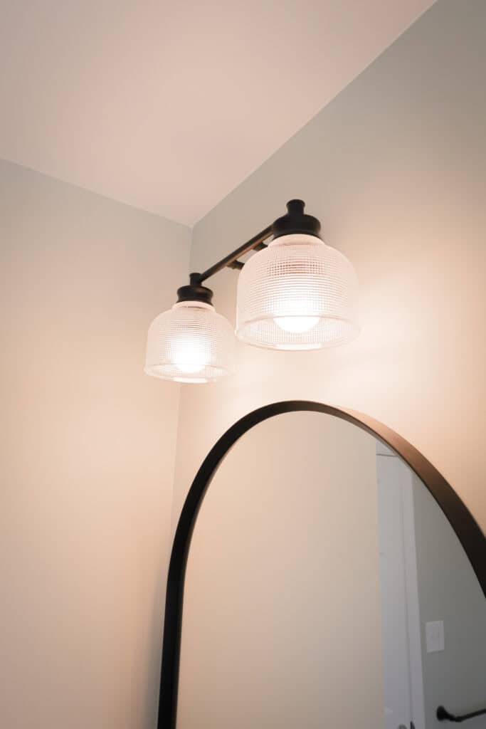 A close-up of a modern bathroom light fixture with two lit glass lamps mounted above a black-framed, arched mirror against a light-colored wall.