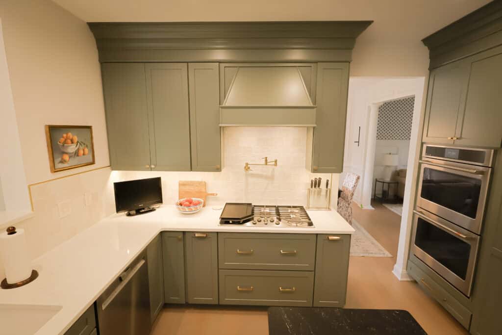 A modern kitchen remodel features green cabinets, white countertops, a gas cooktop, a mounted oven, bowl of red apples, small TV, and fruit painting. Light wood floors and a neutral backsplash complete the refreshed space.