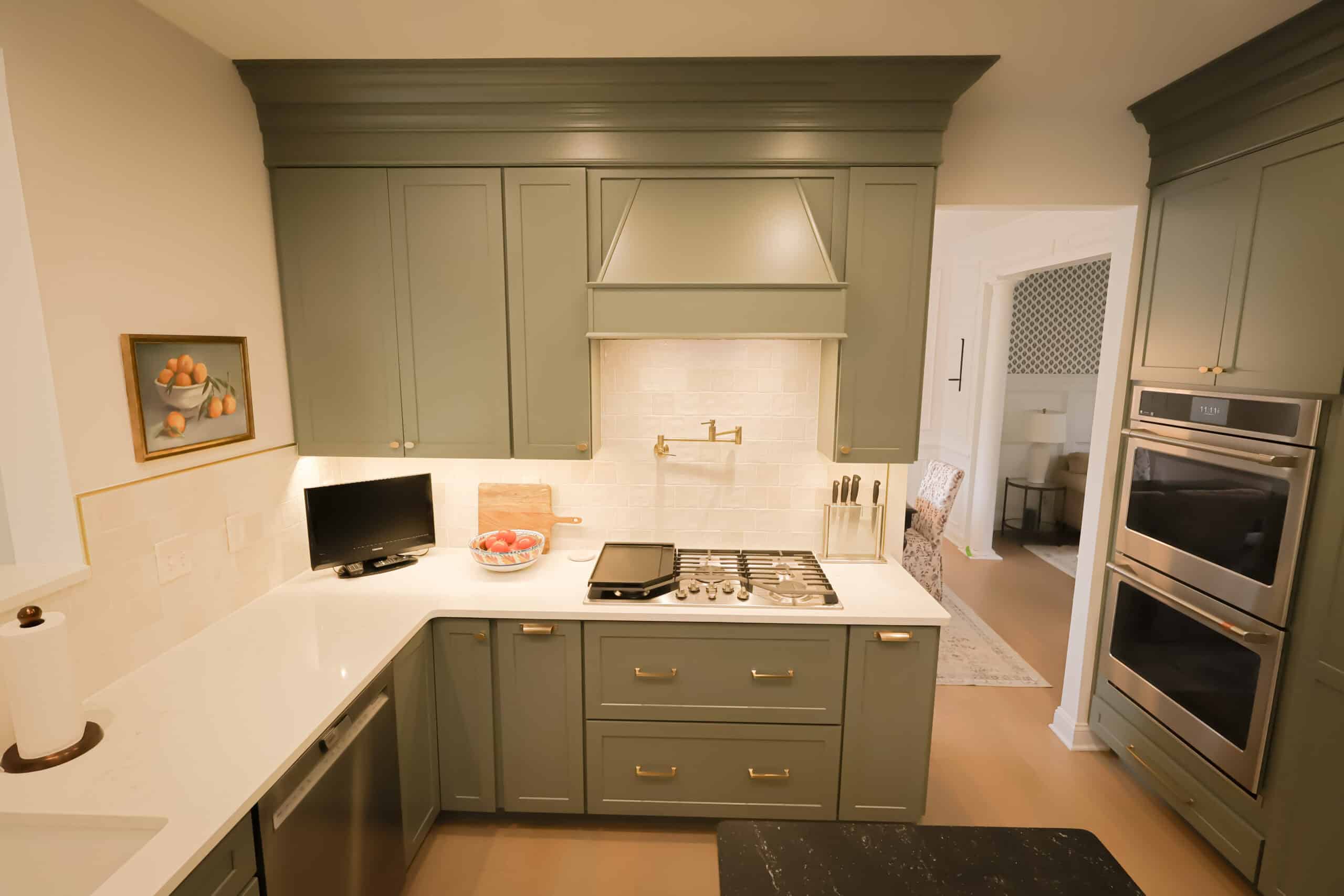 A modern kitchen remodel features green cabinets, white countertops, a gas cooktop, a mounted oven, bowl of red apples, small TV, and fruit painting. Light wood floors and a neutral backsplash complete the refreshed space.