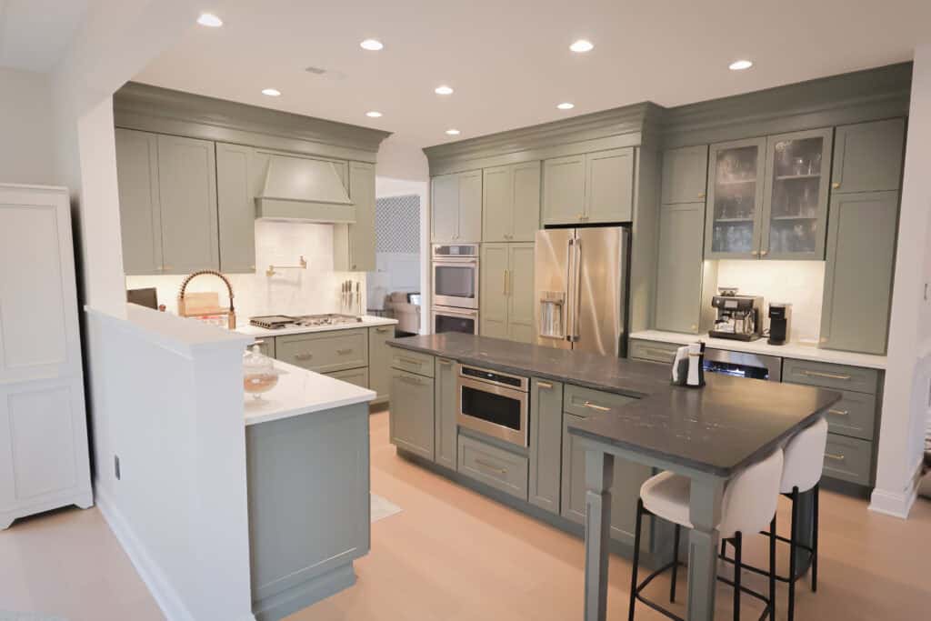A modern kitchen remodel with sage green cabinets, stainless steel appliances, a large island with a black countertop, two white barstools, and under-cabinet lighting creates a bright, clean, and inviting space.