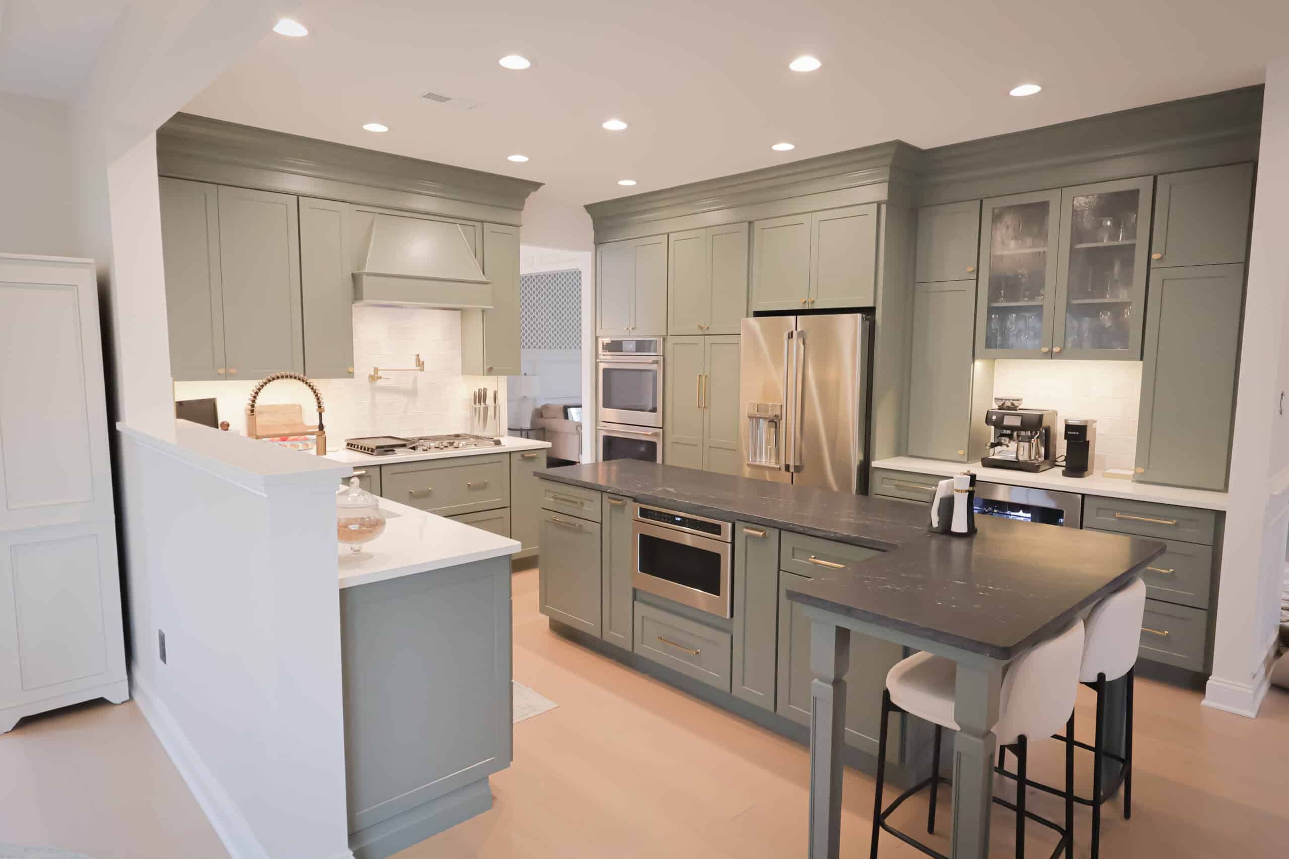 A modern kitchen remodel with sage green cabinets, stainless steel appliances, a large island with a black countertop, two white barstools, and under-cabinet lighting creates a bright, clean, and inviting space.