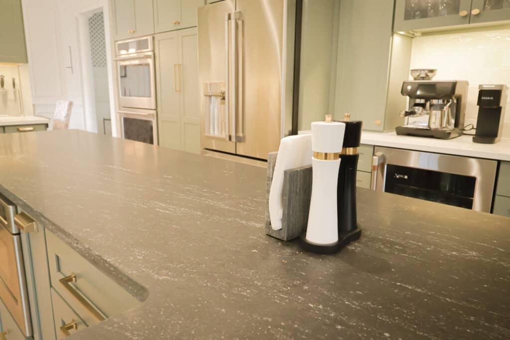 A modern kitchen remodel with green cabinets, stainless steel appliances, and a dark countertop. On the counter sit a salt shaker, pepper shaker, and napkin holder.