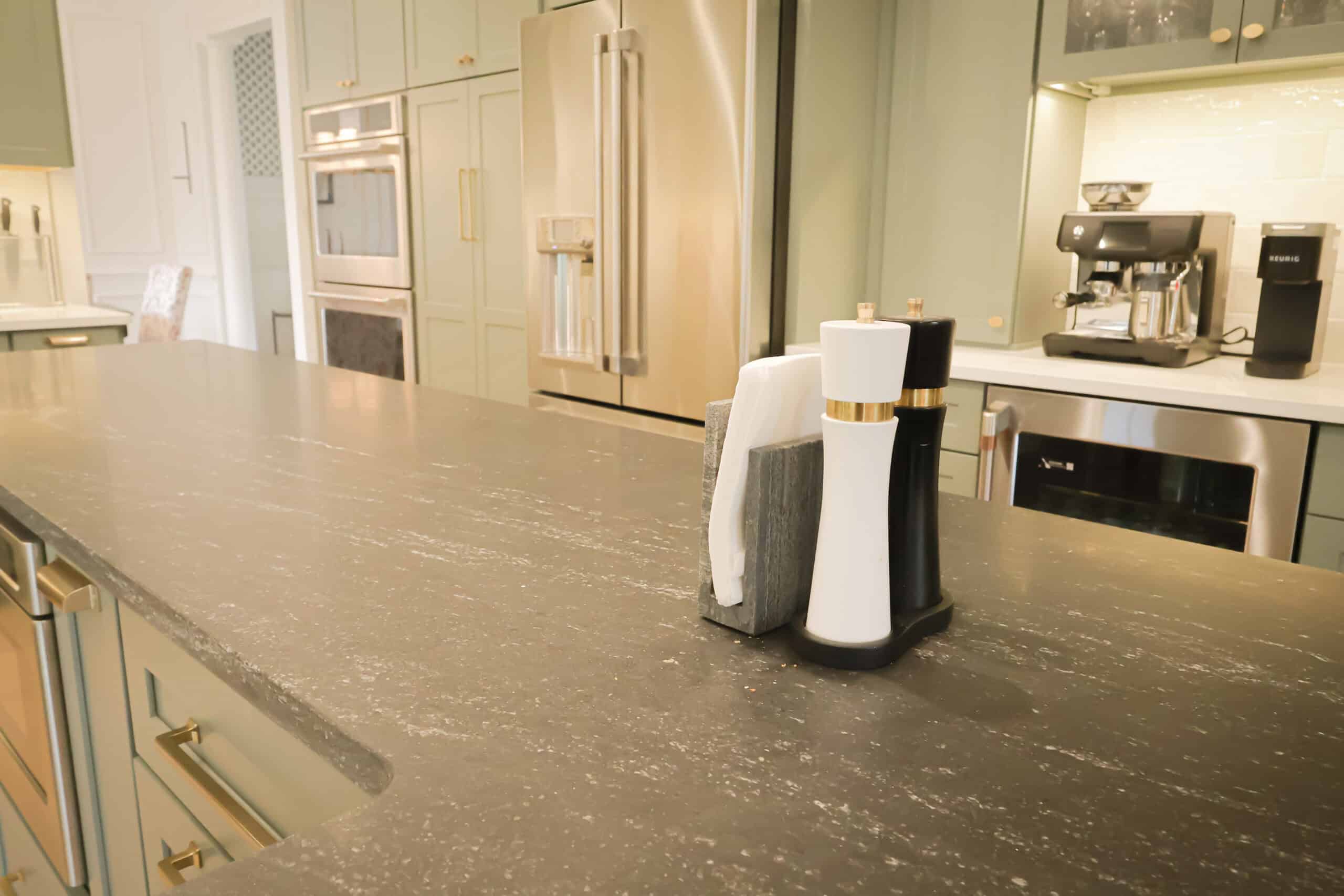 A modern kitchen remodel with green cabinets, stainless steel appliances, and a dark countertop. On the counter sit a salt shaker, pepper shaker, and napkin holder.