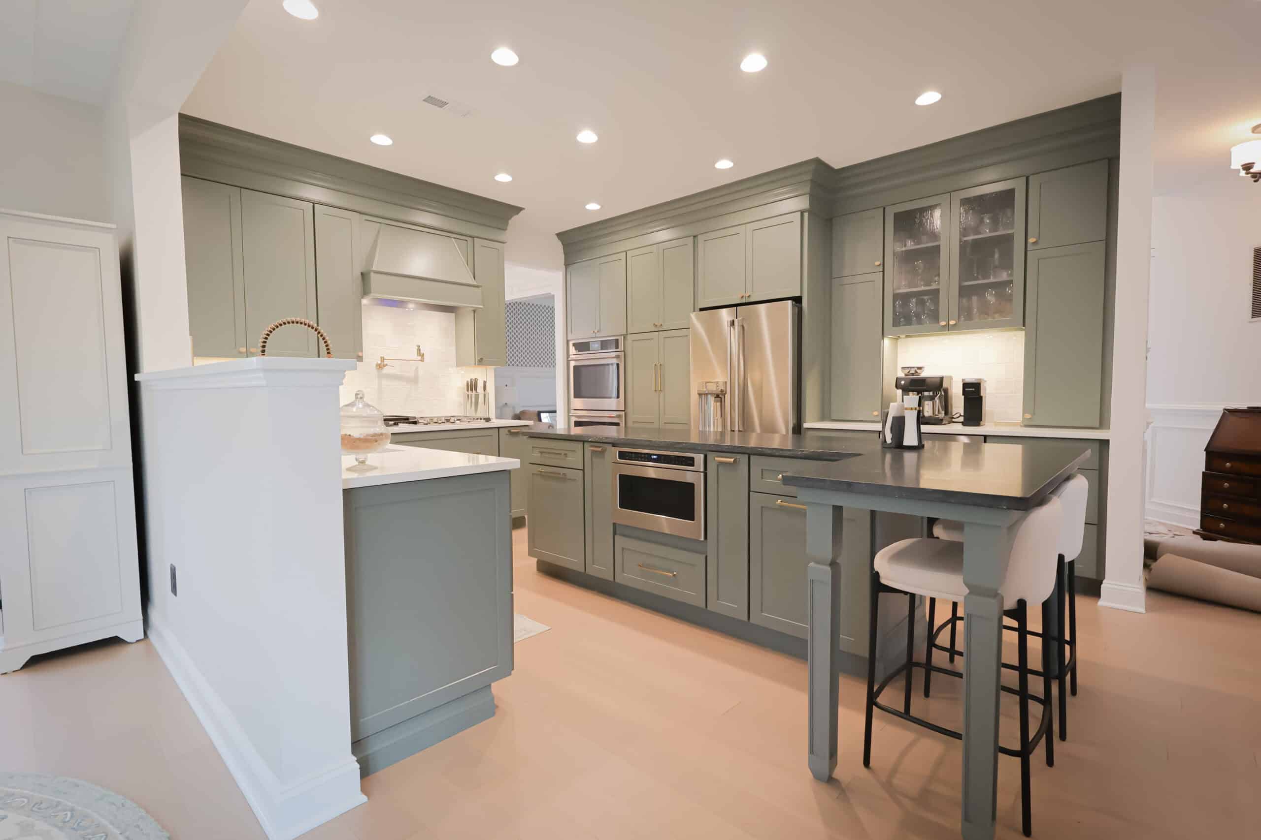 Modern kitchen remodel with sage green cabinets, stainless steel appliances, light wood floors, a center island with a dark countertop, bar stools, and under-cabinet lighting for a bright and elegant atmosphere.