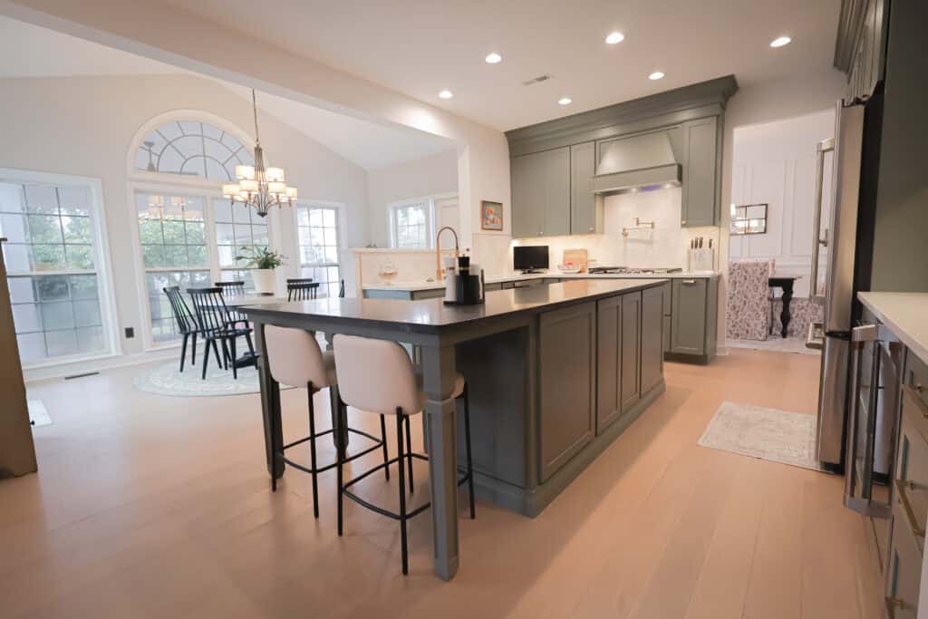Spacious modern kitchen remodel with an island, two upholstered bar stools, green cabinetry, built-in oven, and stove. Dining area with round table and chandelier is visible in the background, while large windows let in natural light.