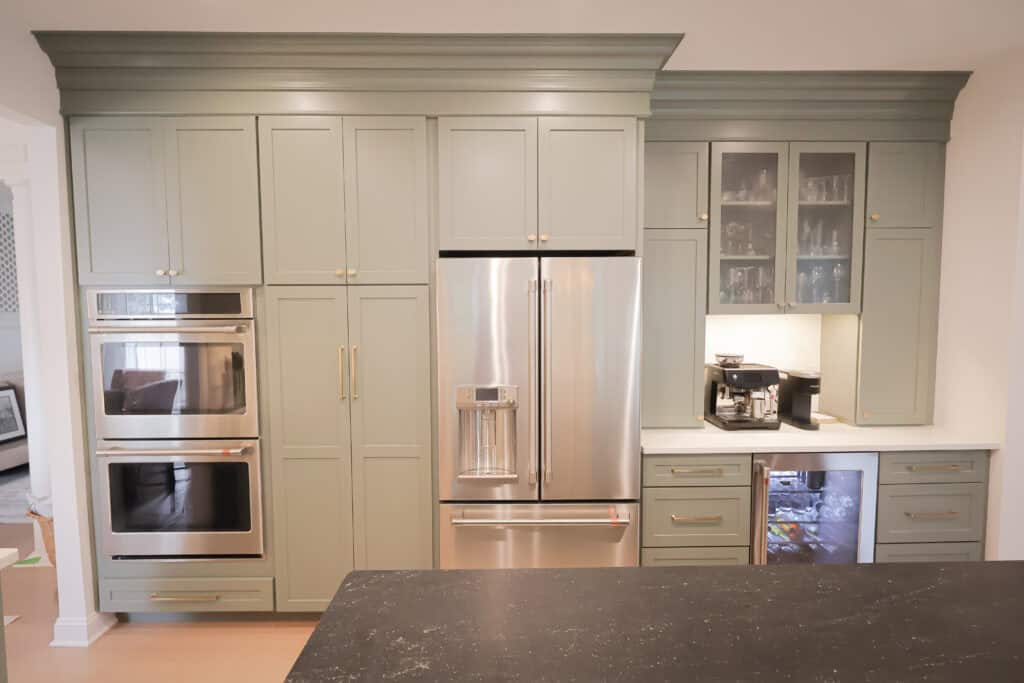 A modern kitchen remodel with sage green cabinets, built-in double ovens, a stainless steel French door refrigerator, a small bar area with glass cabinets, a coffee machine, and a beverage fridge under the counter.