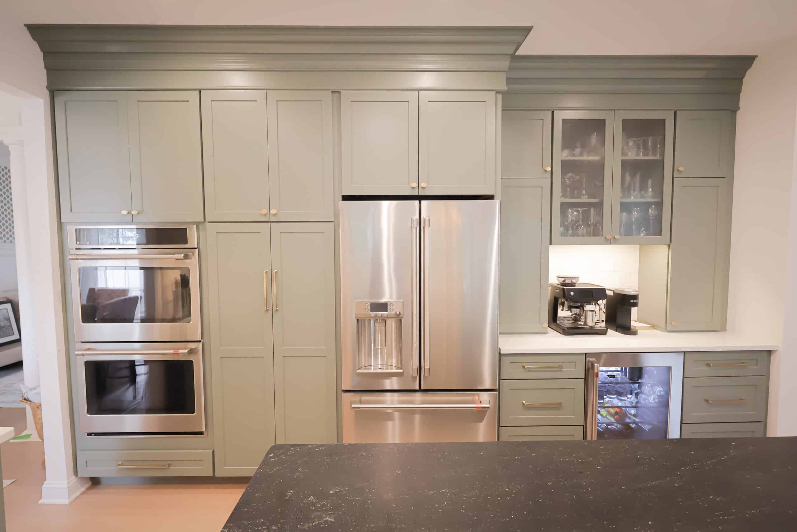 A modern kitchen remodel with sage green cabinets, built-in double ovens, a stainless steel French door refrigerator, a small bar area with glass cabinets, a coffee machine, and a beverage fridge under the counter.