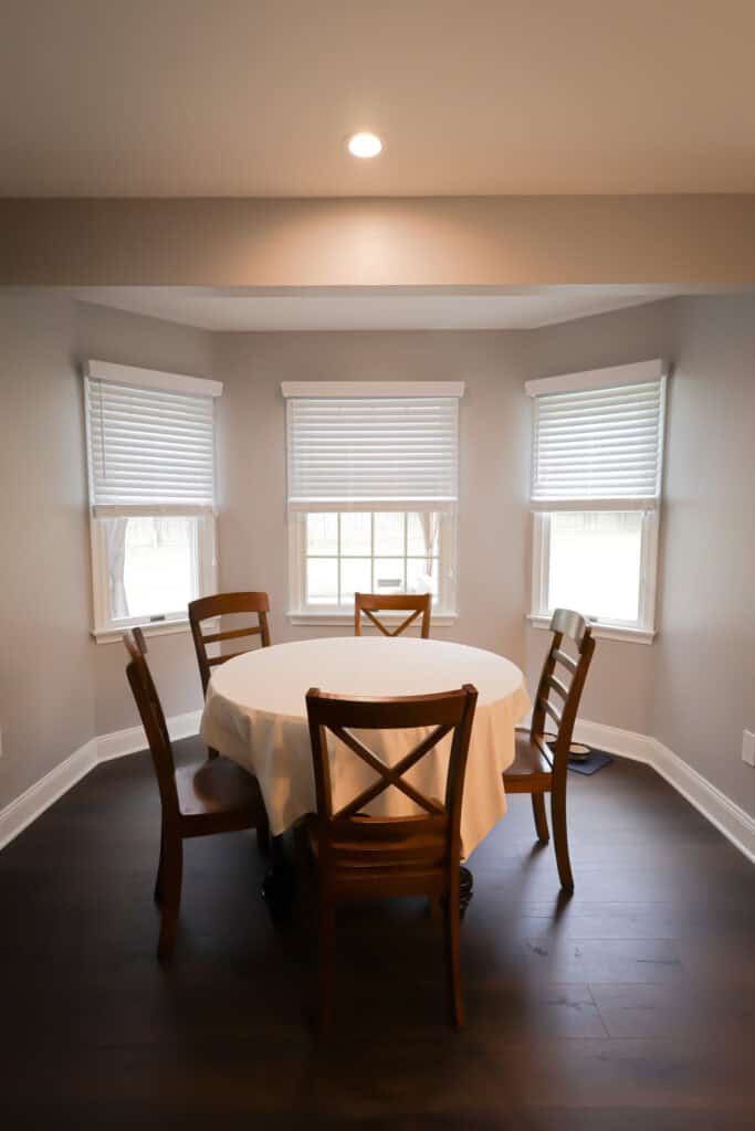 A round table with a white tablecloth and four wooden chairs sits in a corner nook with three windows covered by white blinds, showcasing the elegance often seen in kitchen remodels. The room features dark wood floors and light-colored walls.