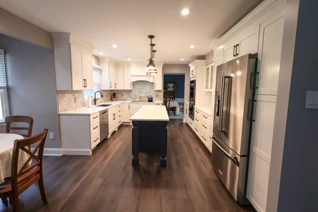 Modern kitchen remodel with white cabinets, stainless steel appliances, a central island featuring a dark base and light countertops, and dark wood floors. A dining table with chairs sits to the left near a window.