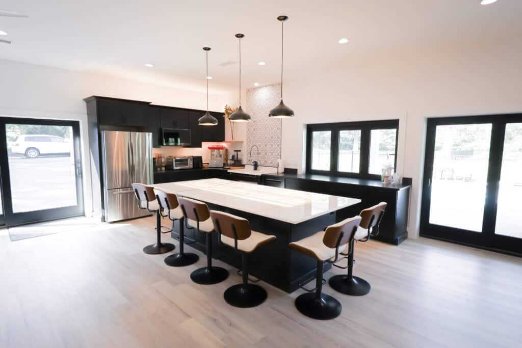 Modern kitchen remodel with a large island, six bar stools, sleek black cabinets, stainless steel appliances, pendant lights overhead, and ample natural light streaming through expansive windows and glass doors.