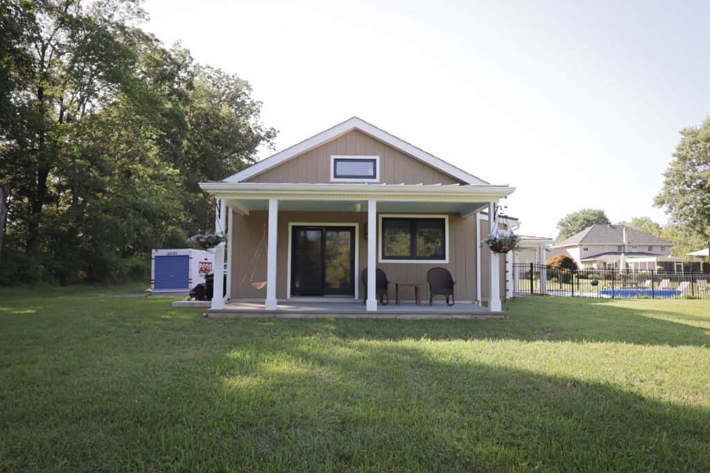 A small beige house with a covered front porch, two chairs, and potted plants sits on a grassy lawn. Trees and other houses are visible in the background under a clear sky.