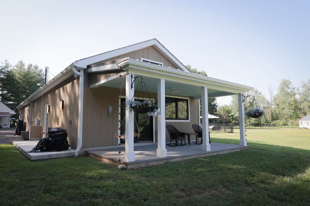 A small tan house with a white-trimmed porch, two chairs, hanging flower baskets, and a swing. The home is surrounded by green grass and trees under a clear sky.