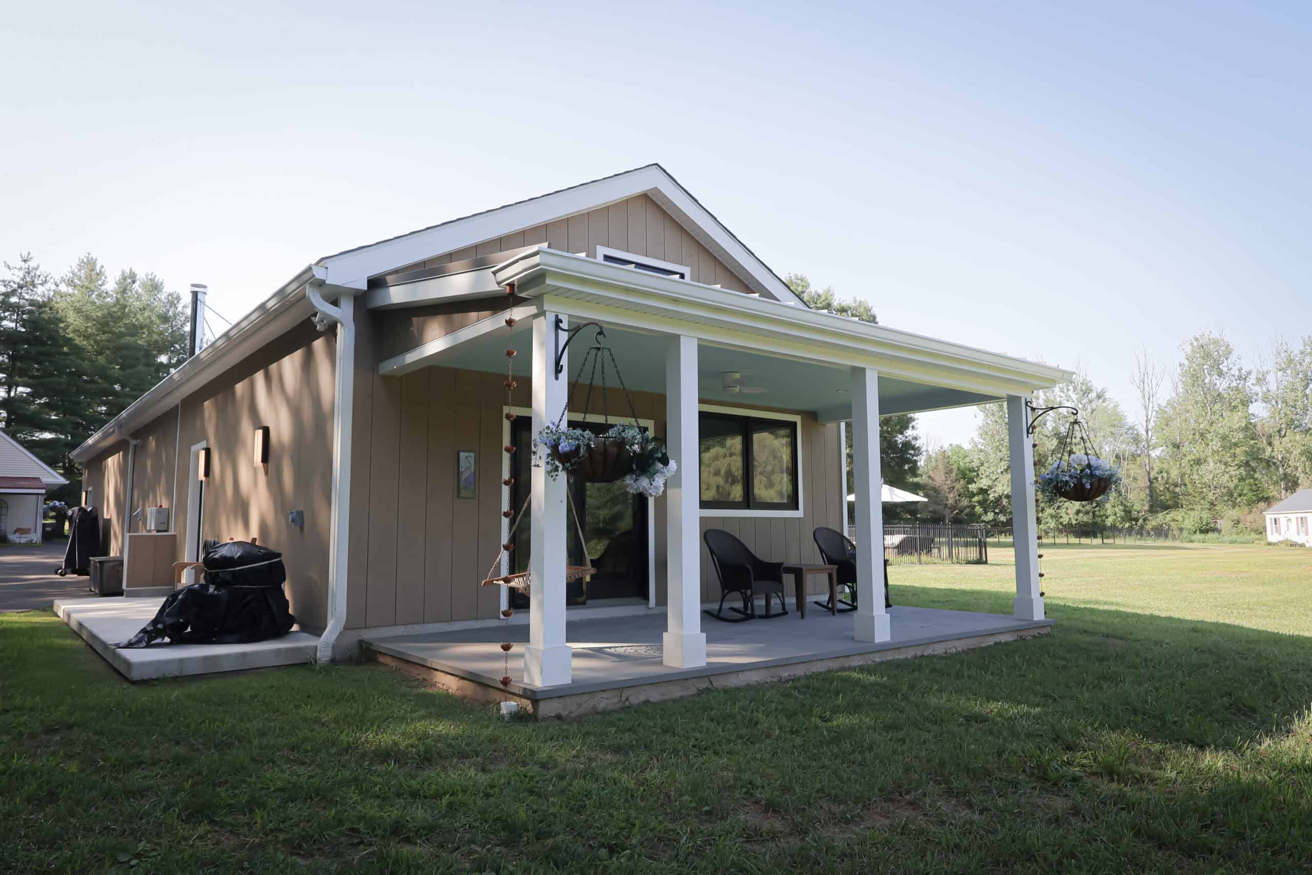 A small tan house with a white-trimmed porch, two chairs, hanging flower baskets, and a swing. The home is surrounded by green grass and trees under a clear sky.