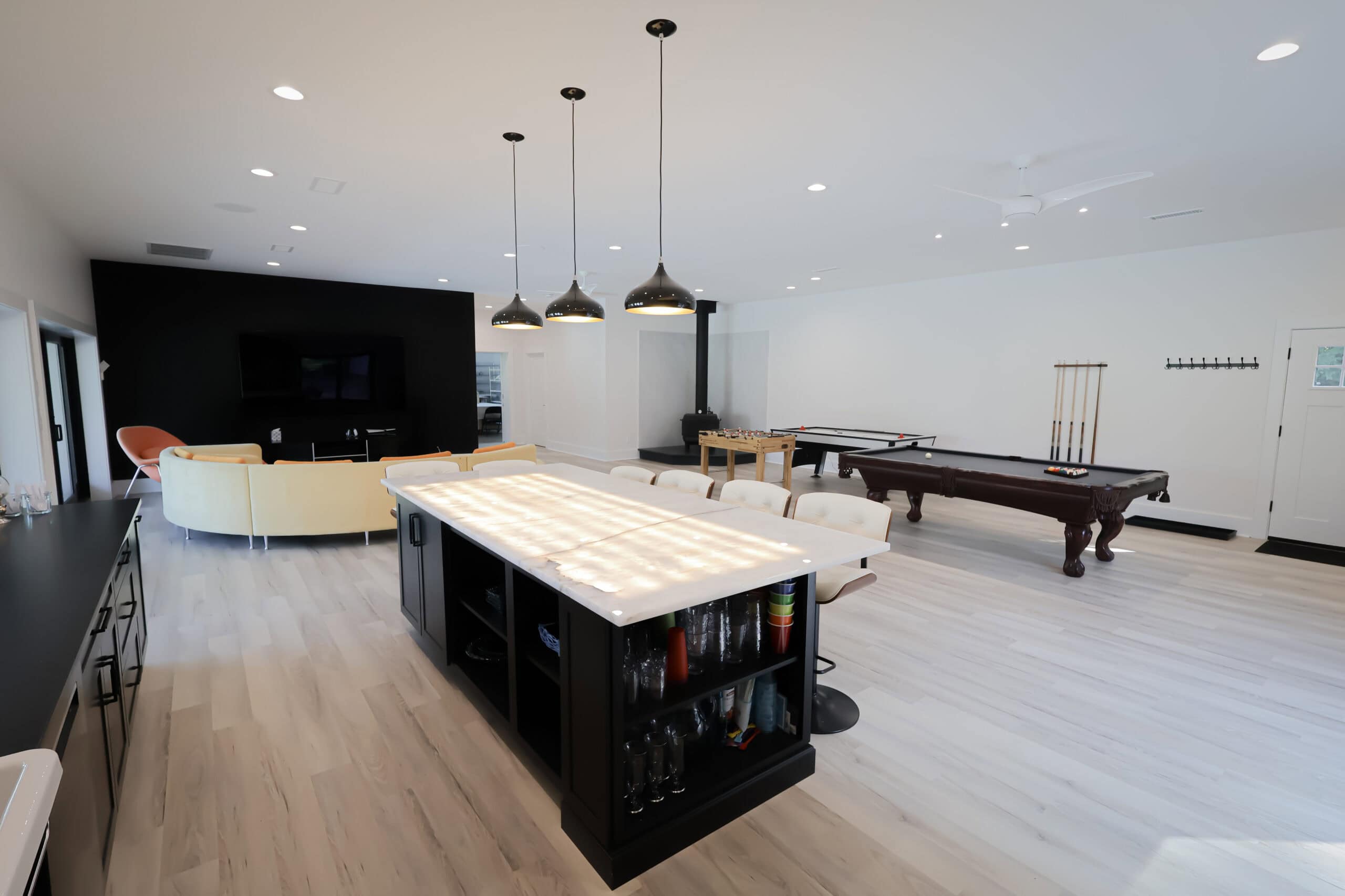 Modern, spacious game room with a kitchen island in the foreground—perfect inspiration for Kitchen Remodels—plus a pool table, lounge area with sofas and TV, pendant lights, light wood floors, and white walls.