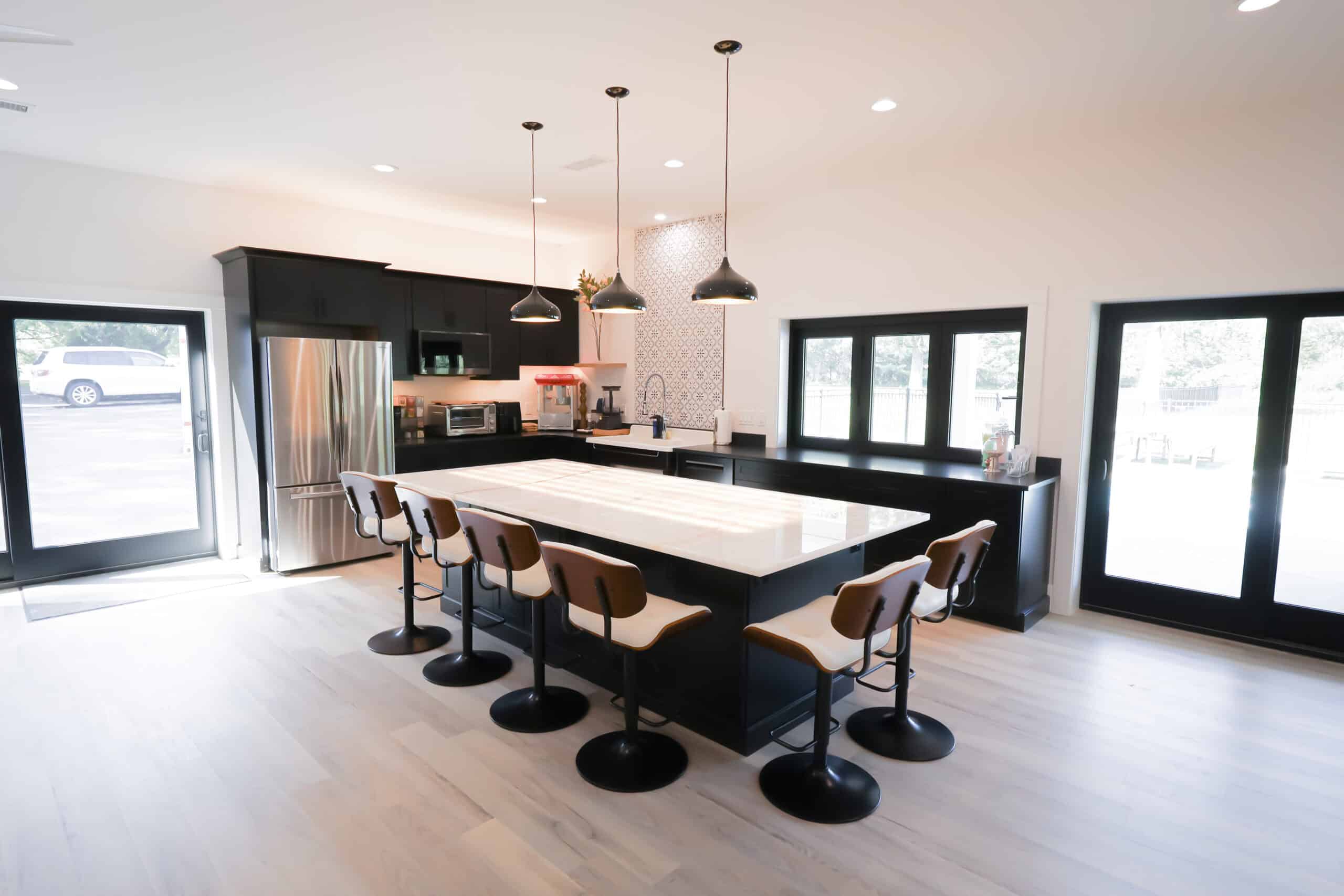 Modern kitchen remodel with a large island, six bar stools, sleek black cabinets, stainless steel appliances, pendant lights overhead, and ample natural light streaming through expansive windows and glass doors.