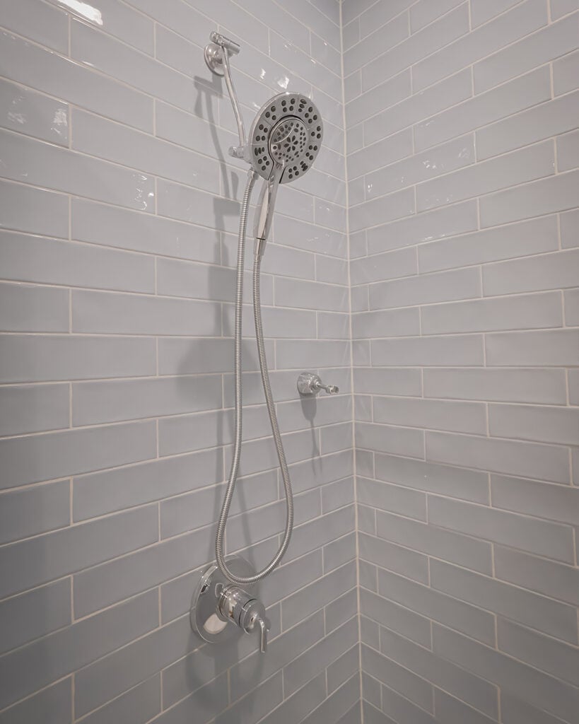 A modern shower with a detachable showerhead and hose is mounted on a wall covered with light gray subway tiles. The fixtures are silver, and there is a control handle below the showerhead.