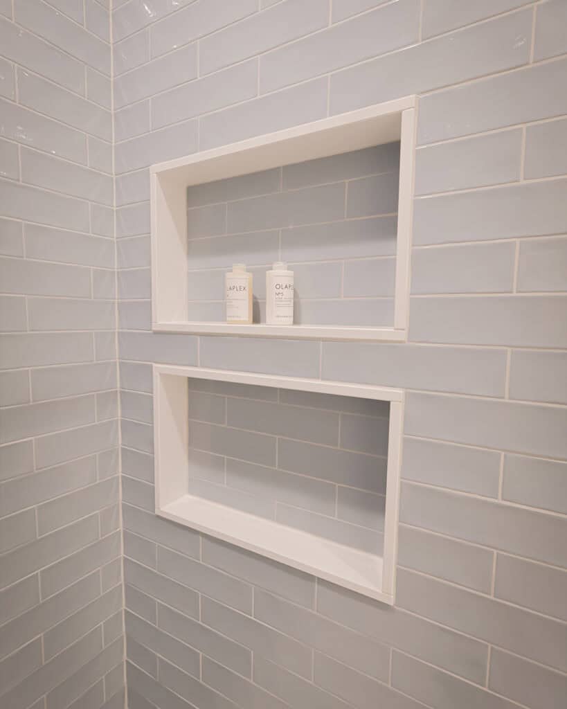 Two white recessed shower shelves set into a wall of light blue rectangular tiles. The upper shelf holds two bottles, while the lower shelf is empty. The scene is clean and minimalistic.