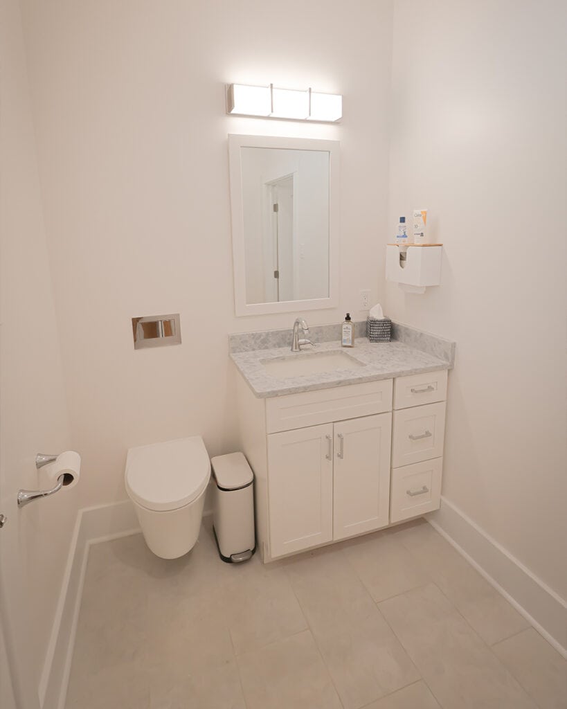 A modern, minimalistic bathroom with a wall-mounted toilet, a white vanity with a marble countertop, a rectangular mirror, and bright overhead lighting. Theres a trash bin beside the vanity and a basket on the counter.