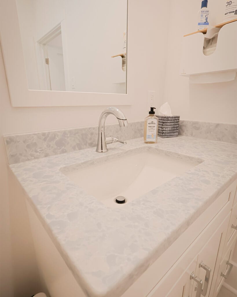 A clean, modern bathroom sink with a silver faucet, marble-patterned countertop, and white cabinets. Soap dispenser, tissue box, and lotion are on the counter; a mirror hangs above the sink.