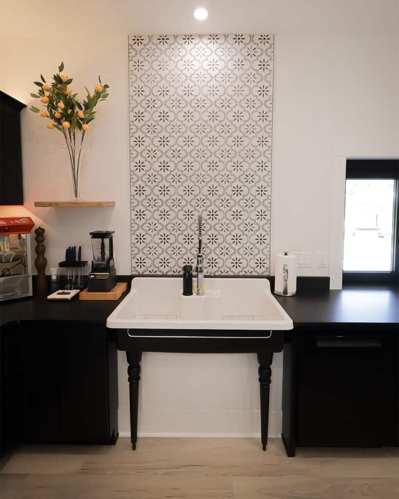 A modern kitchen remodel features a large white farmhouse sink with black legs, a patterned tile backsplash, black countertops, dark cabinets, and a shelf with a vase of green and orange foliage. Natural light enters through a window.