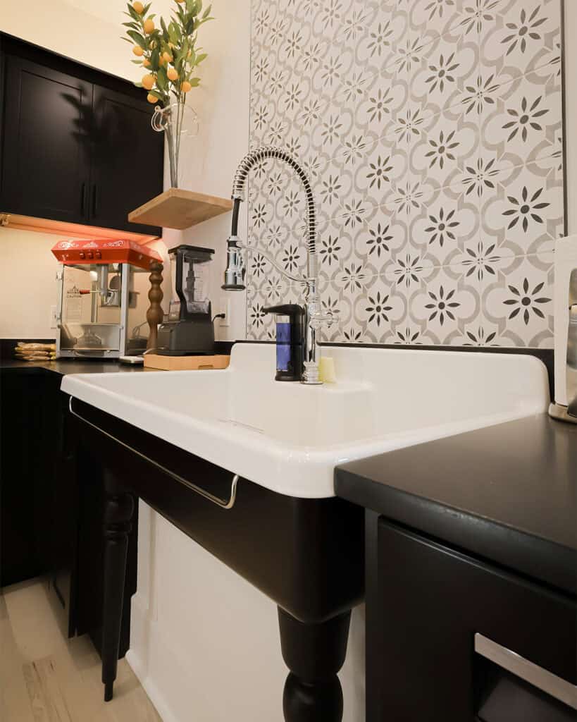 A modern kitchen remodel features a white farmhouse sink with a tall, coiled faucet, black cabinets, patterned tile backsplash, and a popcorn machine on the countertop.