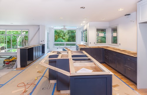 A modern kitchen under construction, featuring dark blue cabinets, countertops not yet installed, large windows, and bright natural lighting. Construction materials and tools are visible throughout the space.