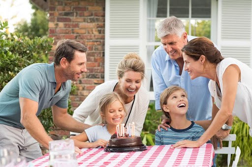 A family gathers outdoors around a table with a birthday cake. Two children sit in front of the cake with lit candles, smiling, while four adults stand behind them, laughing and enjoying the celebration.