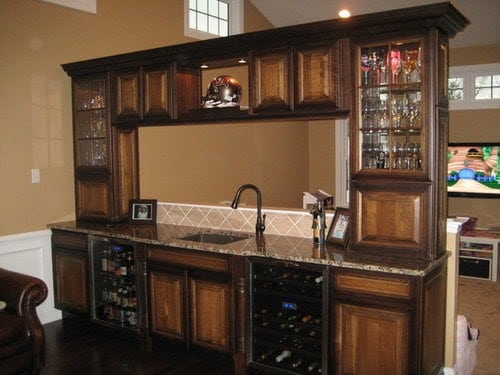 A home bar with dark wood cabinets, a built-in sink, glassware shelves, a wine fridge, and a granite countertop. A TV is visible in the background, and glassware is displayed on the upper shelves.
