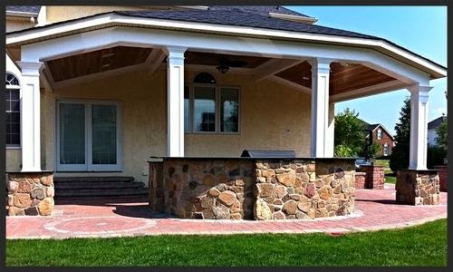 A covered outdoor patio with white columns, stone bar counter, and brick flooring, attached to a house with glass doors and steps leading to the yard.