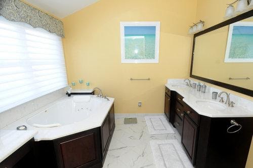 A bright bathroom with yellow walls, a large mirror, dark wood cabinets, a white sink counter, a jacuzzi tub, marble floor tiles, and a window with white blinds.