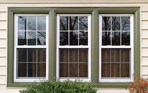A triple window with green trim set in a cream-colored wall. The windows have white frames, vertical and horizontal muntins, and reflect bare tree branches from outside. Shrubs are visible below the windows.