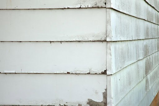 Close-up of the corner of a house with white paint peeling off horizontal wooden siding, revealing patches of bare wood and signs of weathering and age.