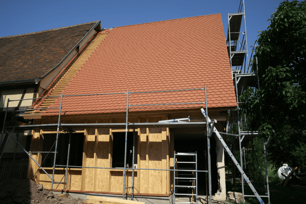 A house under construction, with scaffolding around it. The roof is partially tiled with red shingles, while the exterior walls are unfinished and covered with wooden panels. The sky is clear and blue.