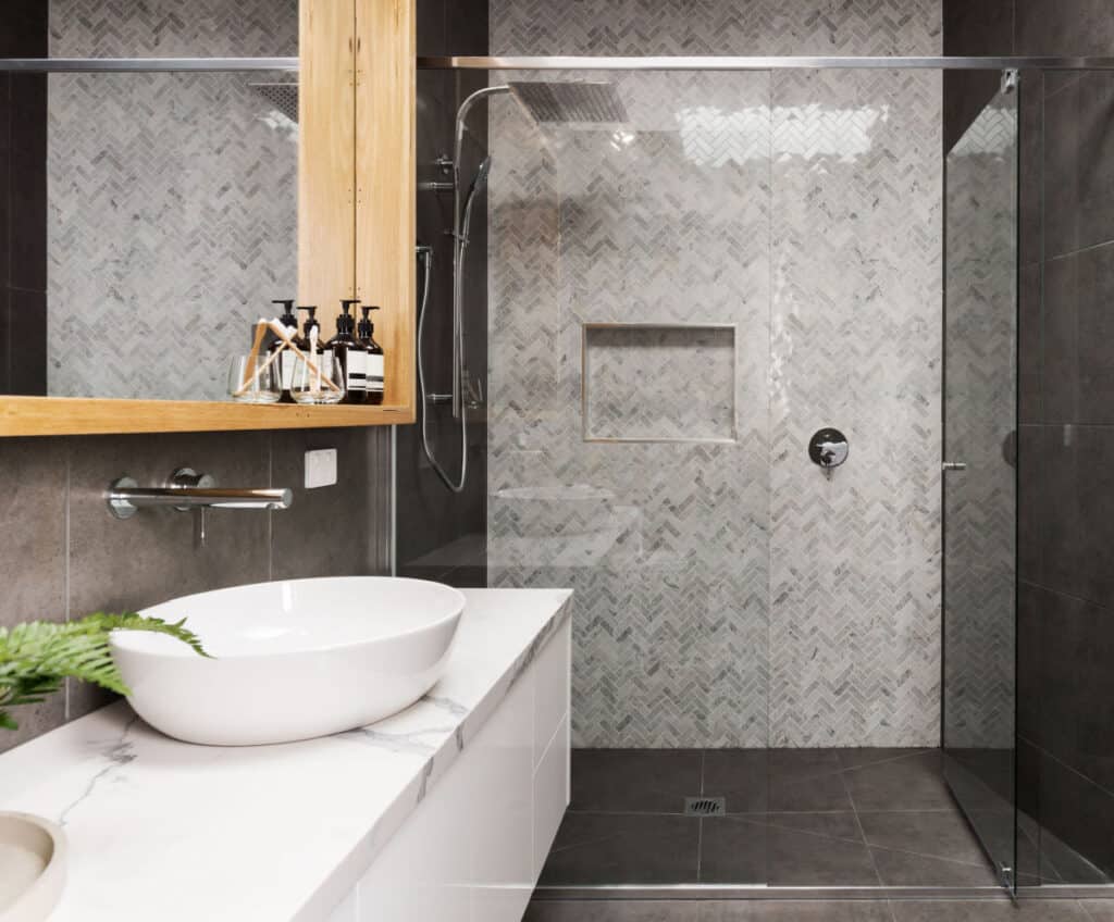 Modern bathroom with a glass-enclosed shower, grey herringbone tile wall, white vessel sink on a marble countertop, wooden-framed mirror, and soap dispensers arranged neatly.