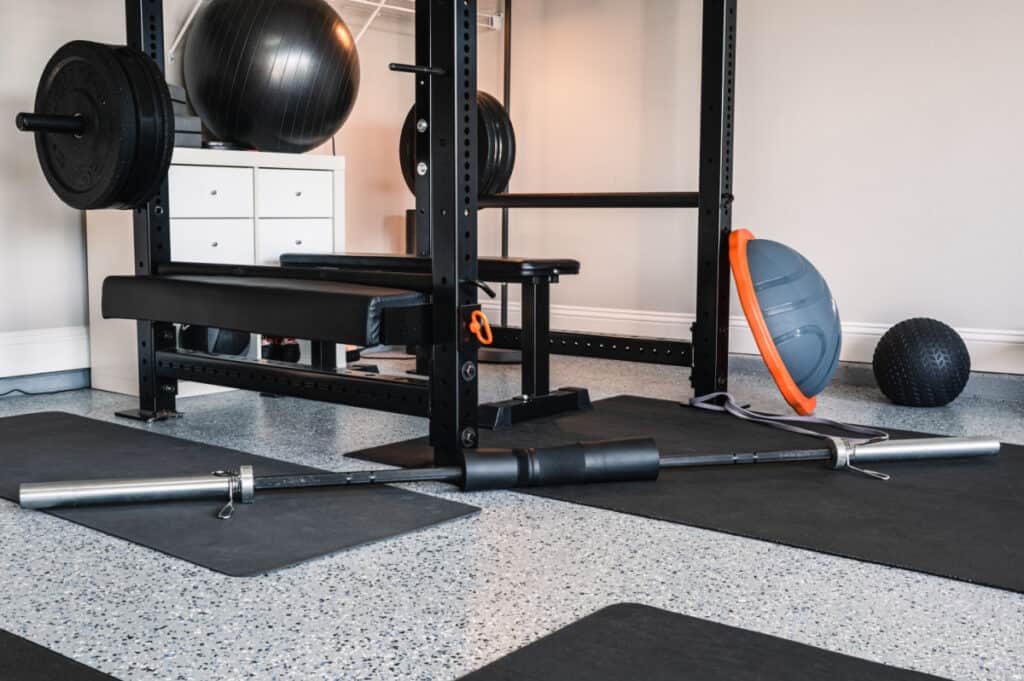 A home gym with a weight bench, squat rack, barbell with weights, a barbell on the floor, exercise balls, a BOSU balance trainer, and black mats on a speckled floor.
