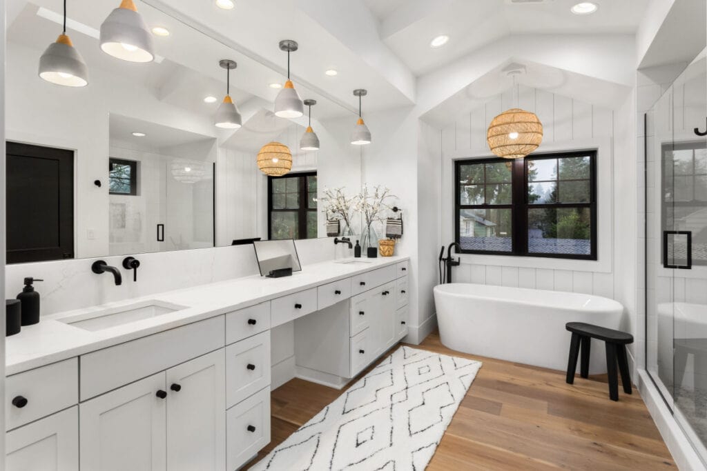 Modern bathroom with white cabinets and countertops, black fixtures, a freestanding tub, a large glass shower, wood flooring, pendant lights, and a geometric white rug. Natural light enters through two black-framed windows.