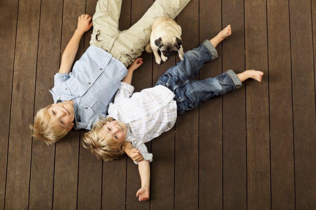 Two young boys are lying on a wooden floor, smiling and looking up. One wears a blue shirt and khaki pants, the other a white shirt and jeans. A small pug puppy sits beside them.