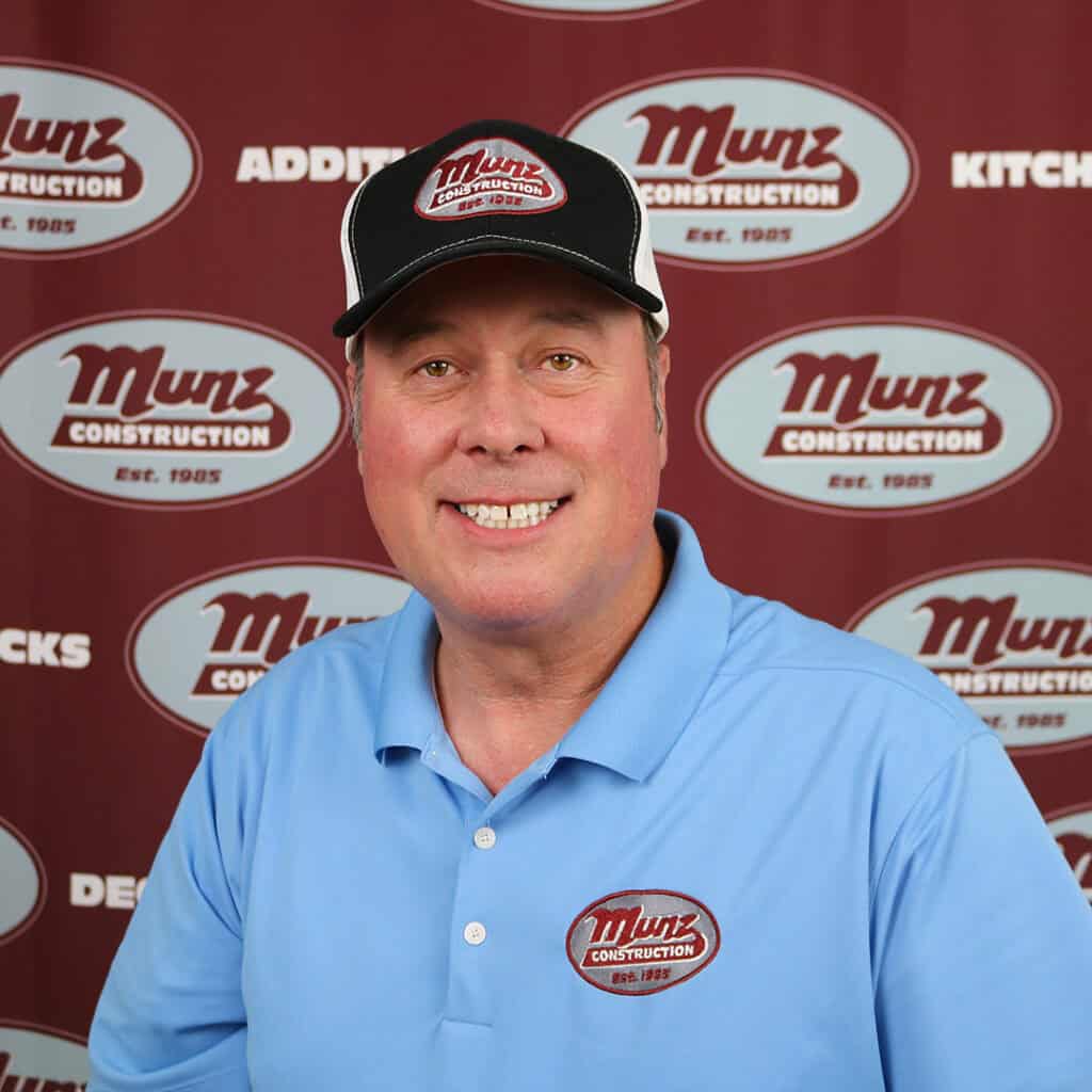 A man in a light blue Munz Construction polo shirt and black cap smiles in front of a Munz Construction-branded backdrop.