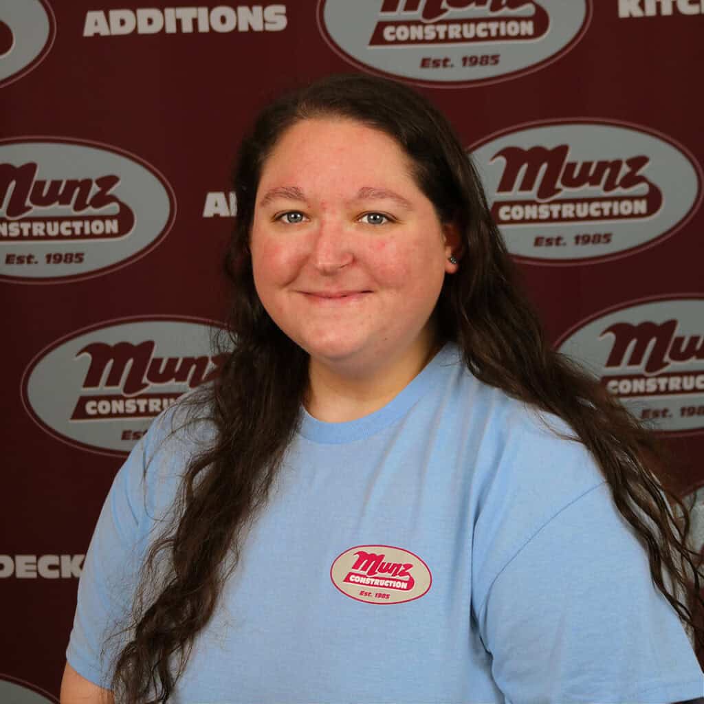 A woman with long brown hair wearing a light blue Munz Construction t-shirt smiles in front of a maroon Munz Construction backdrop featuring the company logo and services.