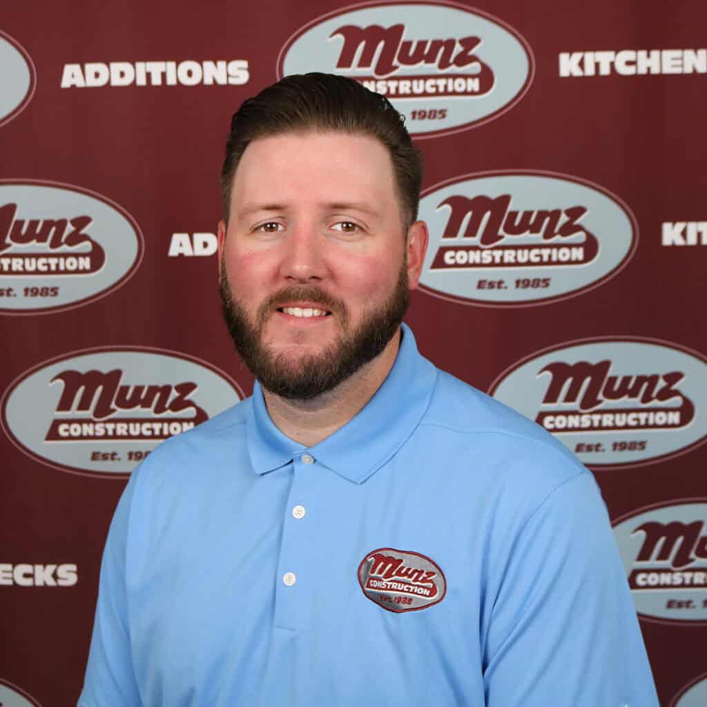 A man with short brown hair and a beard, wearing a light blue polo shirt with a Munz Construction logo, smiles in front of a maroon backdrop with repeated Munz Construction and related text.